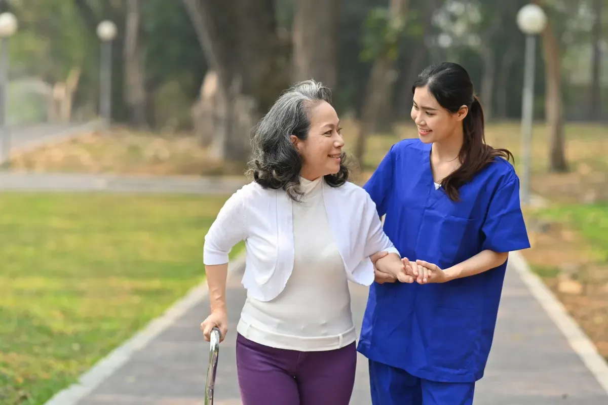 A support worker walking arm in arm with an older woman using a cane through a park